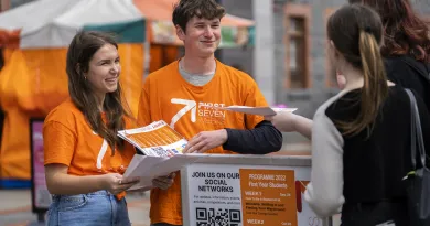 Male and female students wearing orange First 7 Weeks t-shirts speaking to two students, at a First 7 Weeks stand..
