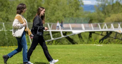 Two female students walking across grass, with the UL Living Bridge in the background, on a sunny day.
