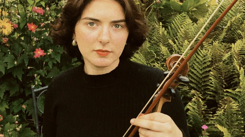 UL graduate Eilidh Pope. She is holding a violin and bow. She is dressed in a black top and is standing in front of green foliage. 