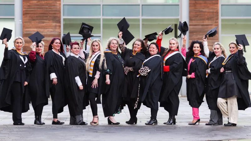A group of 12 Traveller women celebrating their graduation from UL. They are dressed in black graduation robes and are holding their black graduation hats in the air in celebration. 