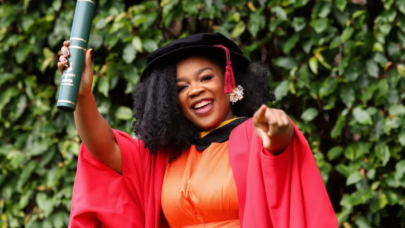 Dr Mamobo Ogoro celebrating her PhD graduation. She is dressed in an orange dress and red graduation robes. She is wearing a black hat with red tassels. She is pointing with one finger and holding a green scroll up in the air with her other hand.