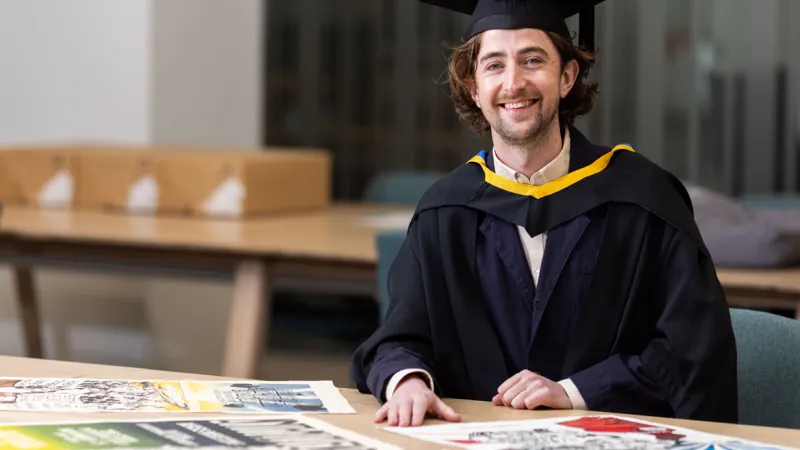 UL graduate Caimin Walsh. He is dressed in a black graduation robe and a black graduation hat. He is seated at a desk and there are screen printed posters on the desk in front of him.
