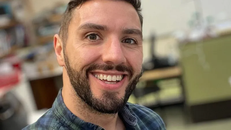 UL graduate Tadgh O'Donovan. He is smiling broadly and is wearing a plaid green and blue shirt. He is standing in a classroom.