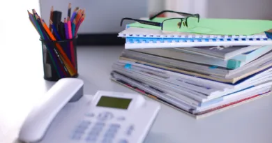 Photo of a landline phone on a table beside a stack of files and a container with different pens and pencils inside.