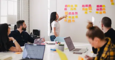 Woman placing sticky notes on a wall as a team of people sitting at a desk with laptops watch her.