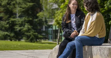 Two girls are sitting outside on a stone bench facing each other chatting. It is a sunny day and there are large trees in the background.