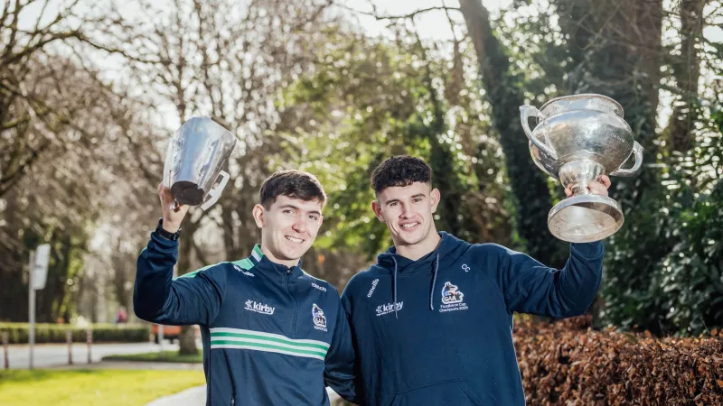 Two young men standing on the grounds of the University of Limerick on a sunny day, smiling and holding up large silver trophies. They are wearing navy athletic tops with sports logos, standing on a tree-lined path with greenery and hedges in the background.