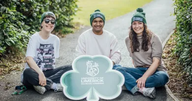 3 students wearing UL green beanies sitting on the ground with a shamrock sign with the UL logo on it