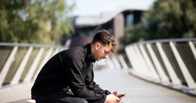 A man is sitting outside on a bench looking at his phone. In the background there are trees and a large building visible